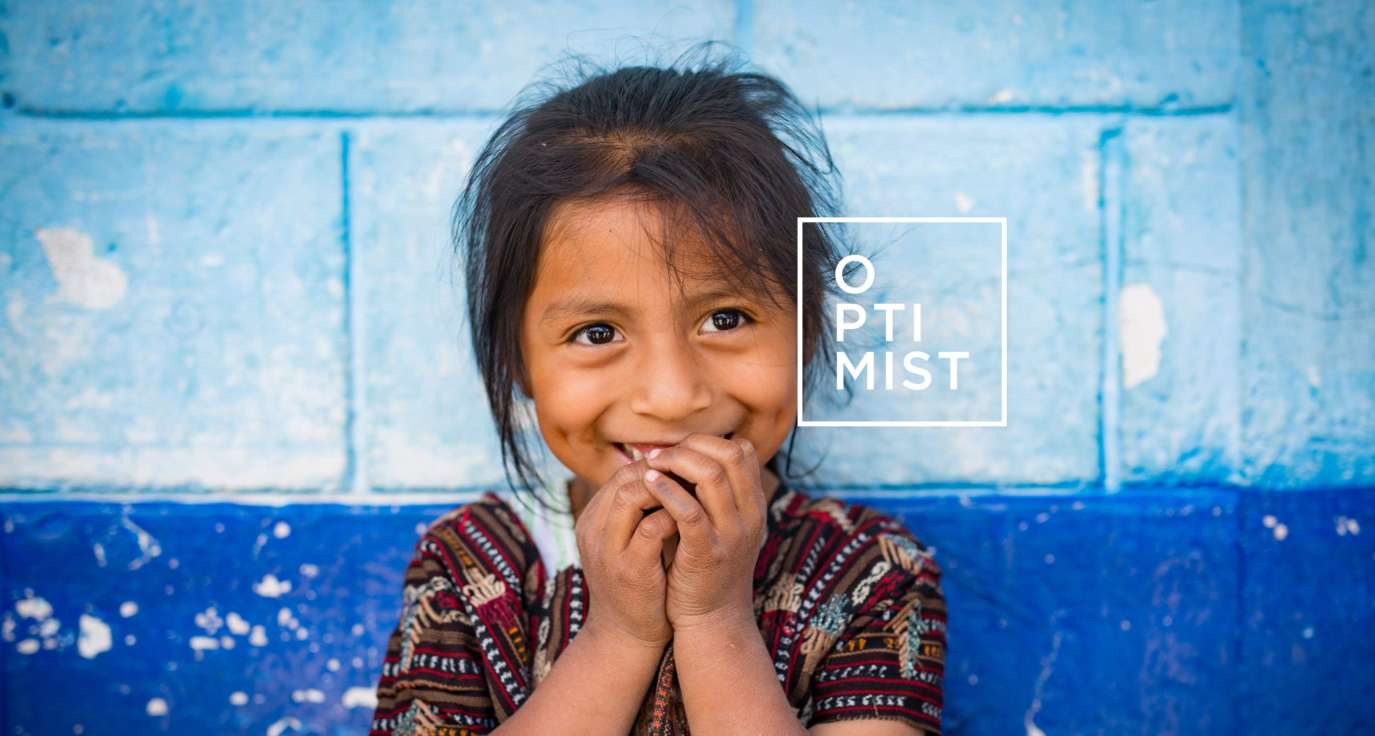 honduran girl smiling with hands in mouth in front of blue wall with optimist logo on image