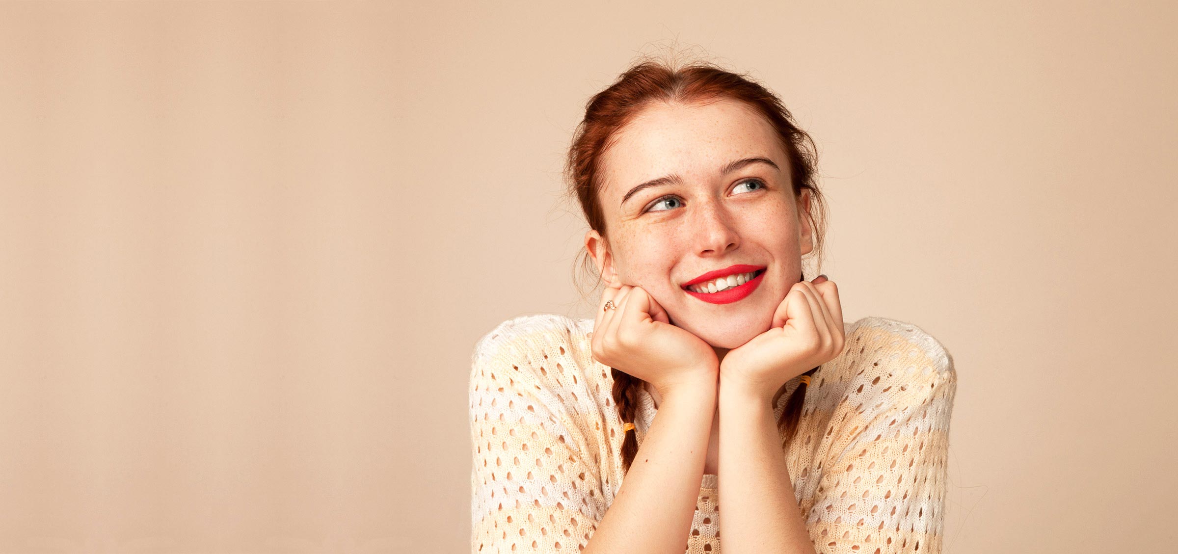 teenage girl with red lipstick and red hair smiling with head in hands on peach backdrop